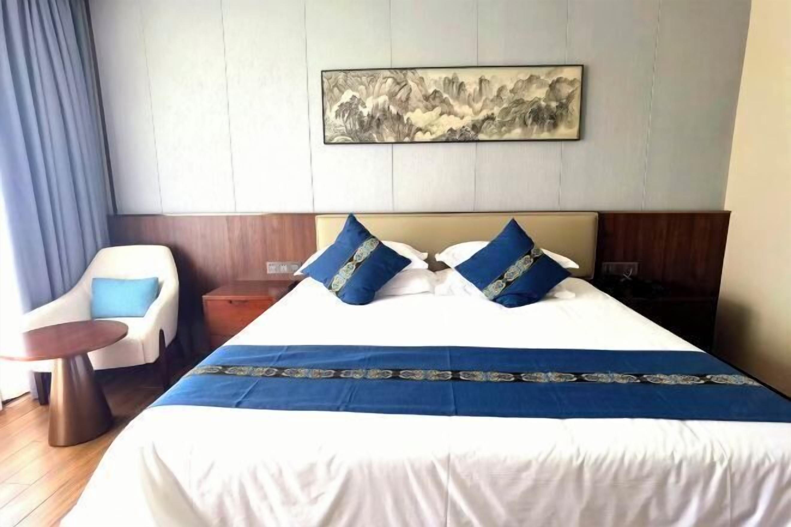 A hotel room with a large bed, two pillows, a blue bedspread, a chair, a small table, and a painting on the wall.