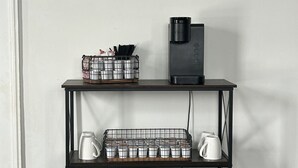 Un comptoir à café avec une enseigne encadrée, une machine à café Keurig, un panier en métal contenant des ustensiles, un plateau en métal avec des tasses à café et deux plantes en pot.