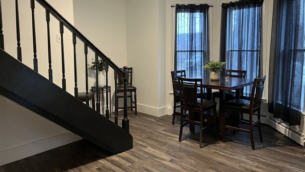 Un coin repas avec une table et des chaises en bois, un escalier et de grandes fenêtres avec des rideaux.