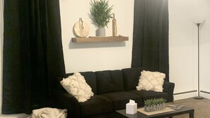Un salon avec un canapé marron foncé, deux coussins blancs et moelleux, une petite table d’appoint avec une boîte de mouchoirs et une plante, un panier contenant un tissu blanc et une lampe de sol noire.