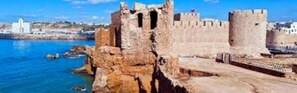 Una fortificación costera con muros y torres de piedra, con vistas a una masa de agua y un paisaje urbano al fondo.