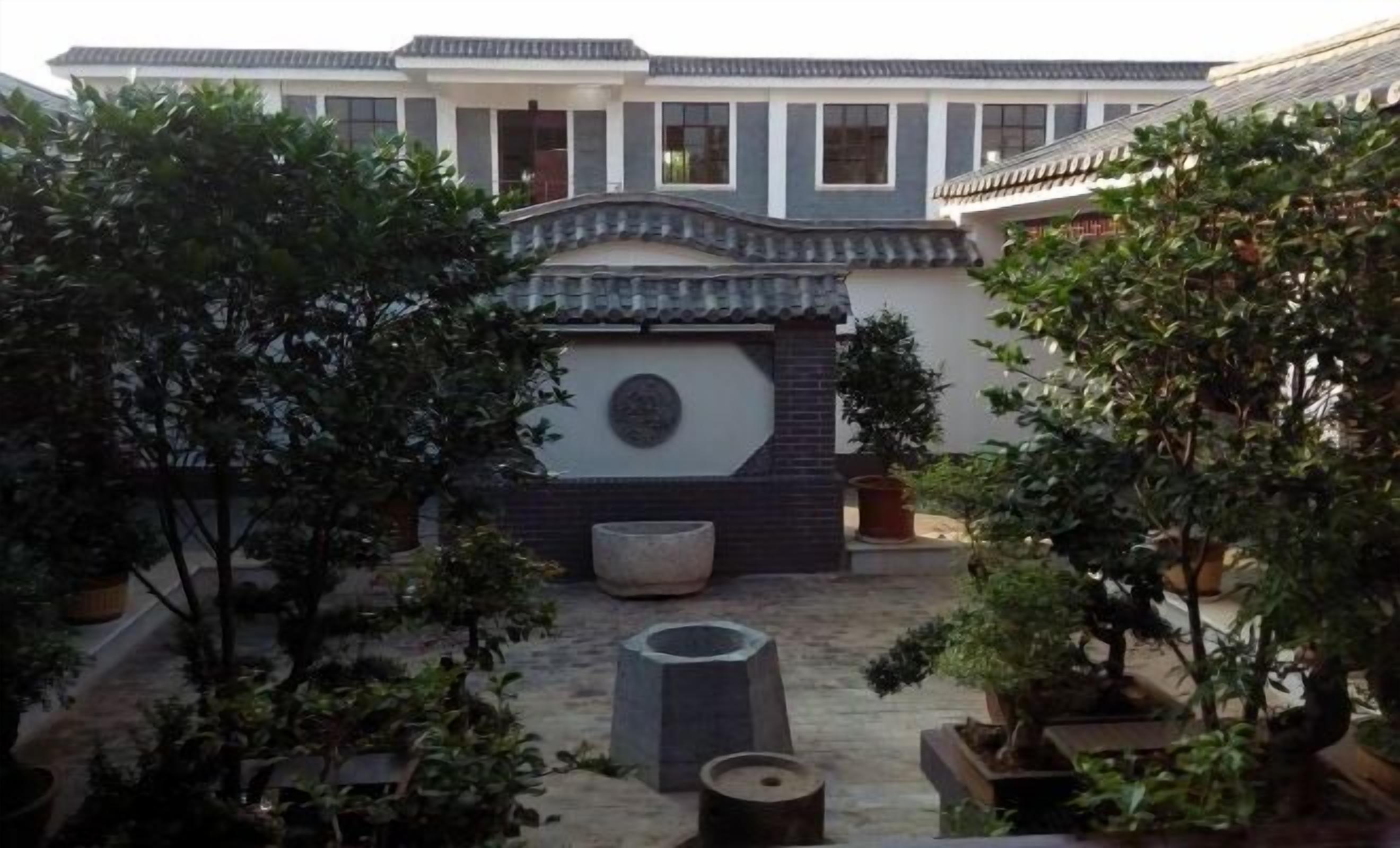 A courtyard with traditional architecture, including a tiled roof and a stone wall with a circular emblem. There are potted plants and a stone bench.
