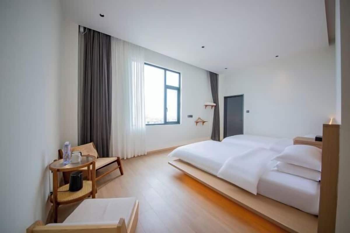 A modern hotel room with a large bed, a small desk with a chair, a window with curtains, and a door.