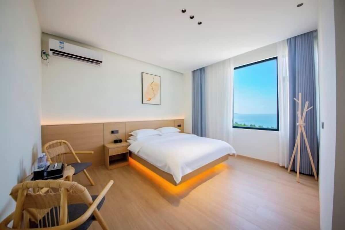 A modern hotel room with a bed, bedside table, chair, and a large window with a view.