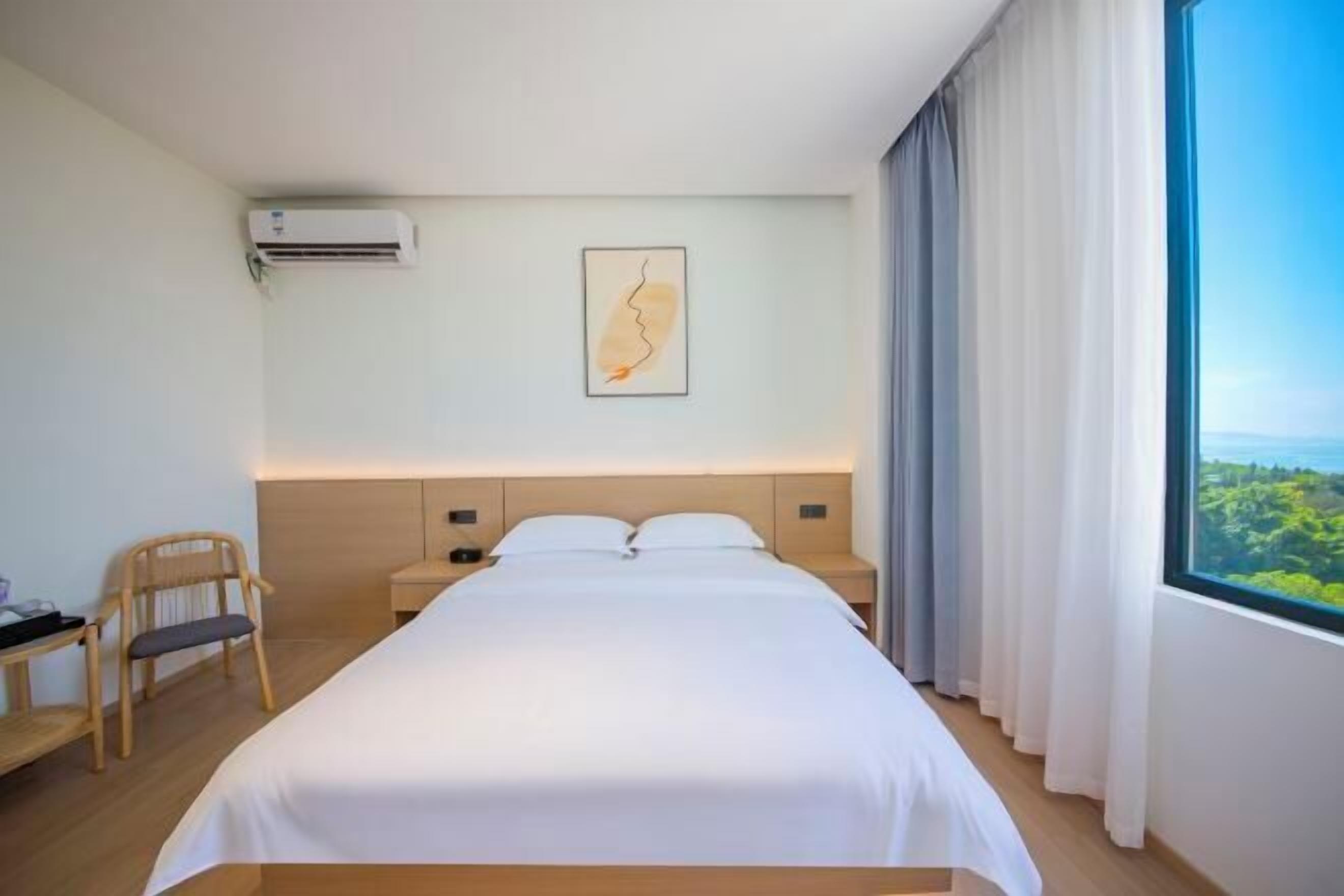 A hotel room with a bed, a chair, a desk, an air conditioner, a framed picture, and a window with curtains.