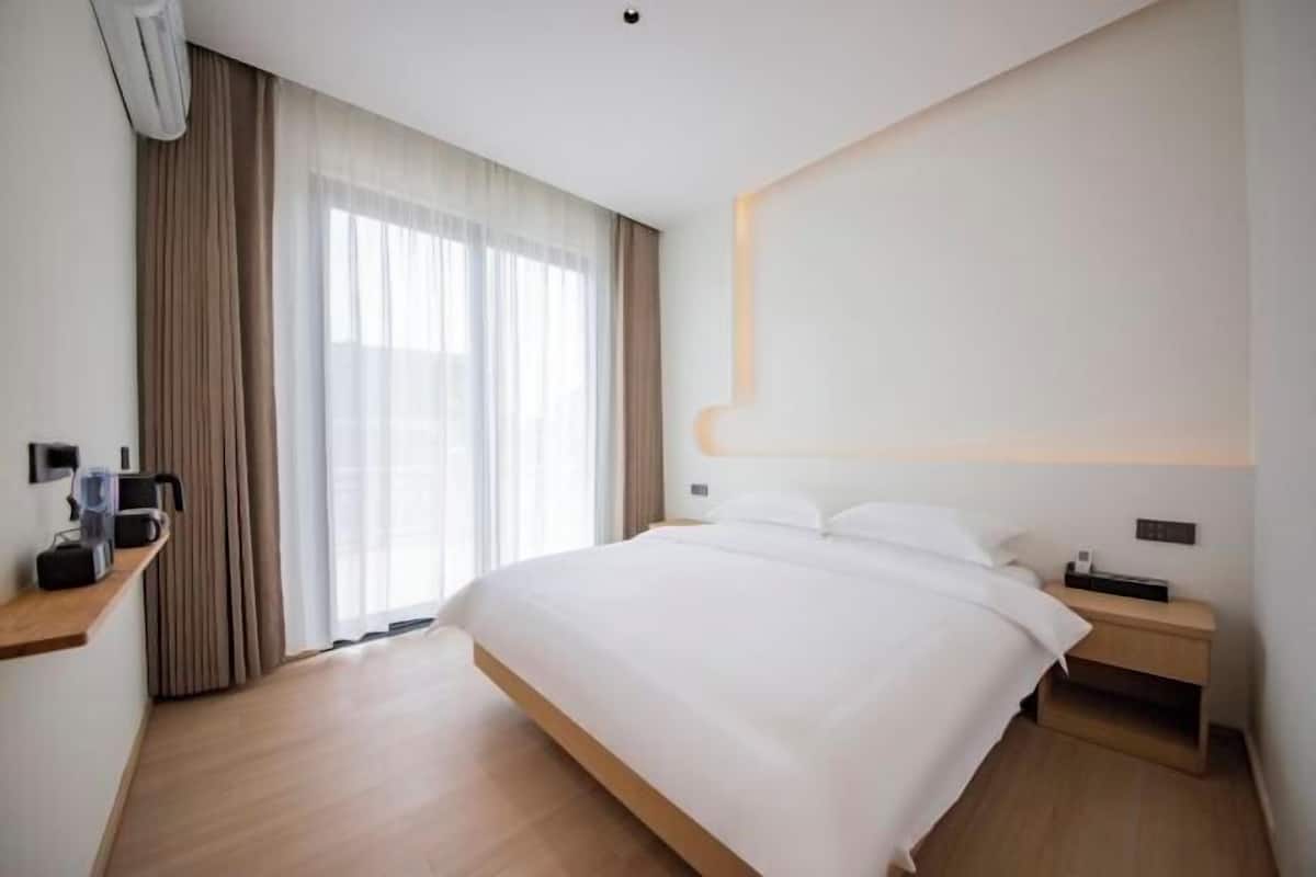 A modern hotel room with a large bed, a bedside table, a nightstand, a wall-mounted light, and a window with curtains.