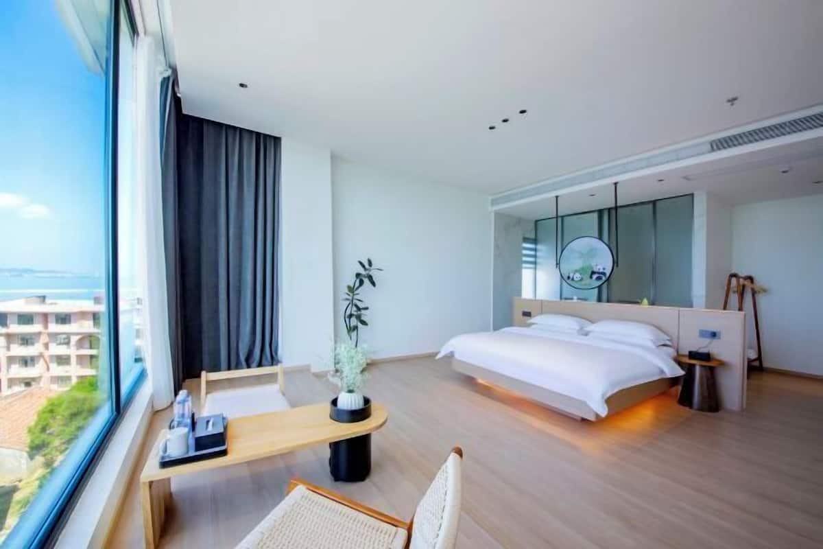 A modern hotel room with a large bed, a desk with a chair, a small table, a potted plant, a mirror, and a view of the city.