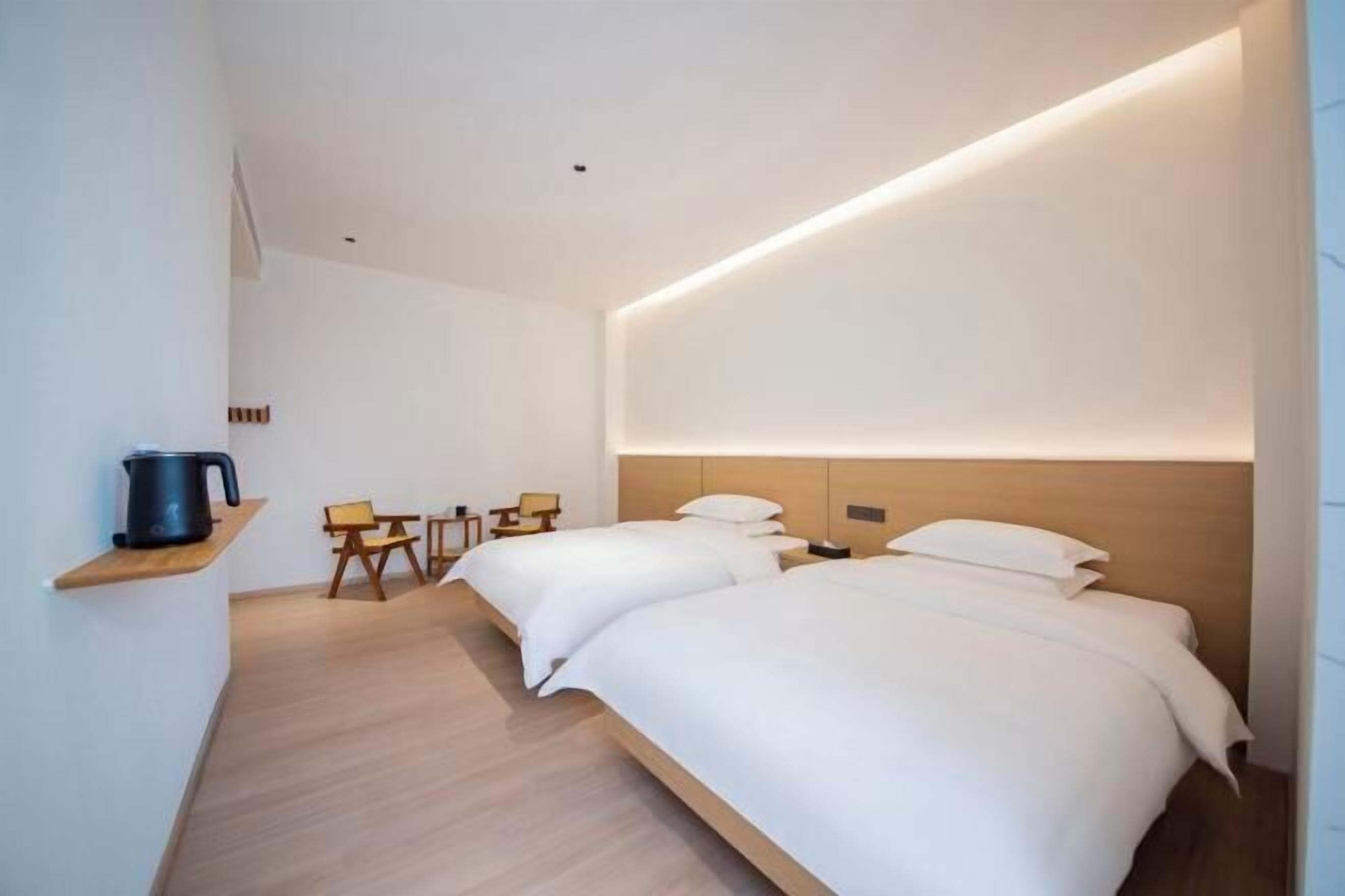 Una habitación de hotel moderna con dos camas, un cabecero de madera, una mesita con sillas y un hervidor en un estante.