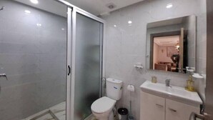 Moderni kylpyhuone, jossa on lasinen suihkuseinä, wc, peilillä varustettu pesuallas ja roskakori.