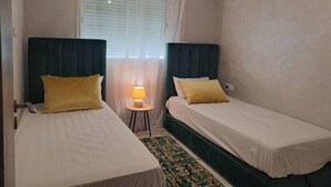 Hotellihuone, jossa on kaksi erillistä sänkyä, pieni pöytä lampun kanssa ja ikkuna, jossa on sälekaihtimet.
