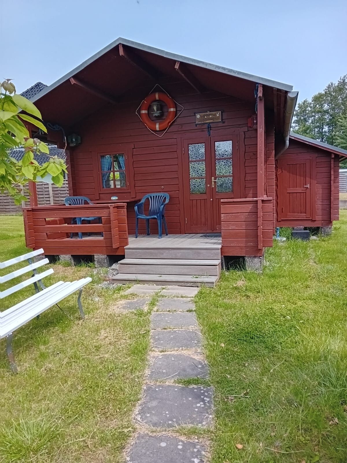 A red wooden cabin with a porch, two blue plastic chairs, a white bench, and a stone pathway leading to the entrance.