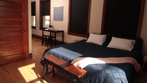 A bedroom with a wooden door, a desk with a chair, a bed with a blue quilt, and a bench.