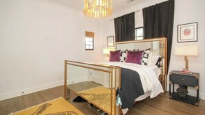 A bedroom with a bed, a nightstand, a lamp, a window with curtains, and a chandelier.