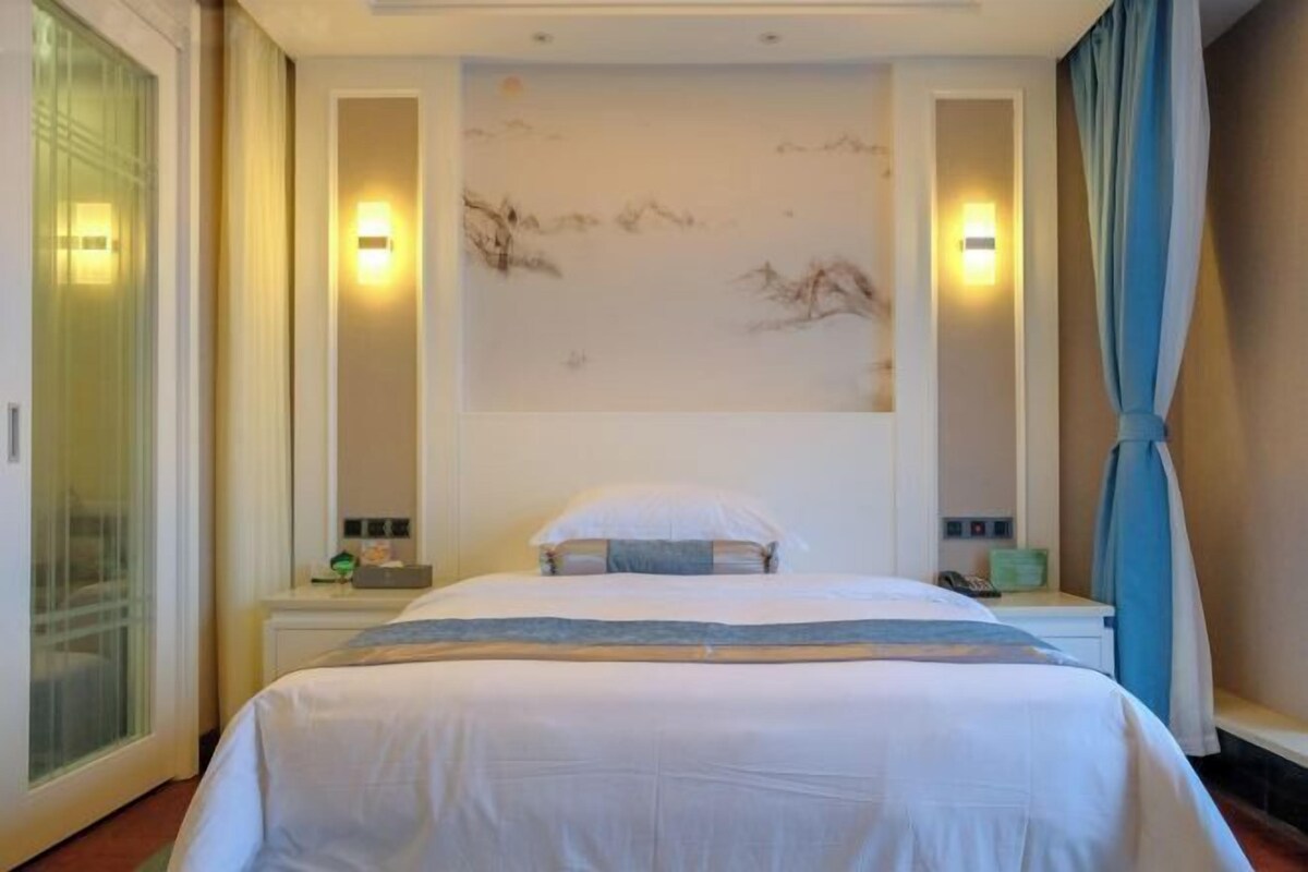 Une chambre d’hôtel avec un lit, des tables de chevet, une œuvre d’art accrochée au mur et une fenêtre avec des rideaux.