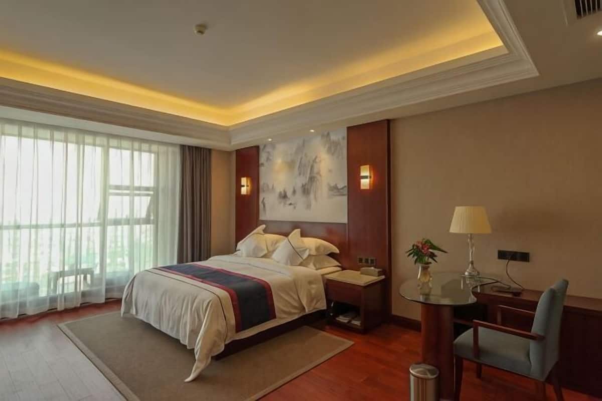 Una habitación de hotel con cama, mesita de noche, lámpara, sillón y un ventanal grande.