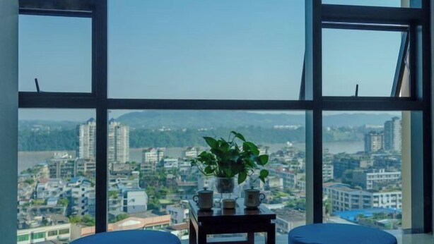 A room with a large window, a small table with a plant, and two blue cushioned seating areas. - Jiu Long Ying Bin Hotel (Yibin)