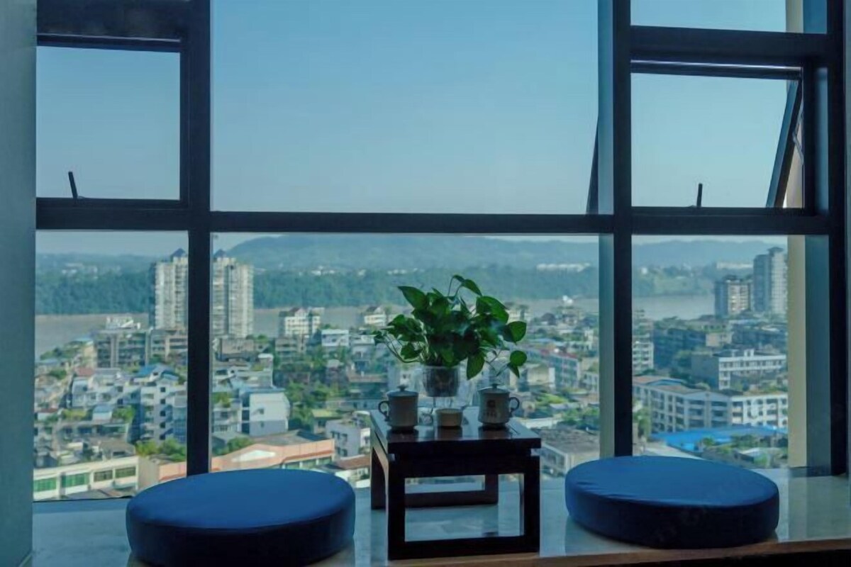 Une pièce dotée d’une grande fenêtre, d’une petite table ornée d’une plante et de deux coins salon bleus avec des coussins.