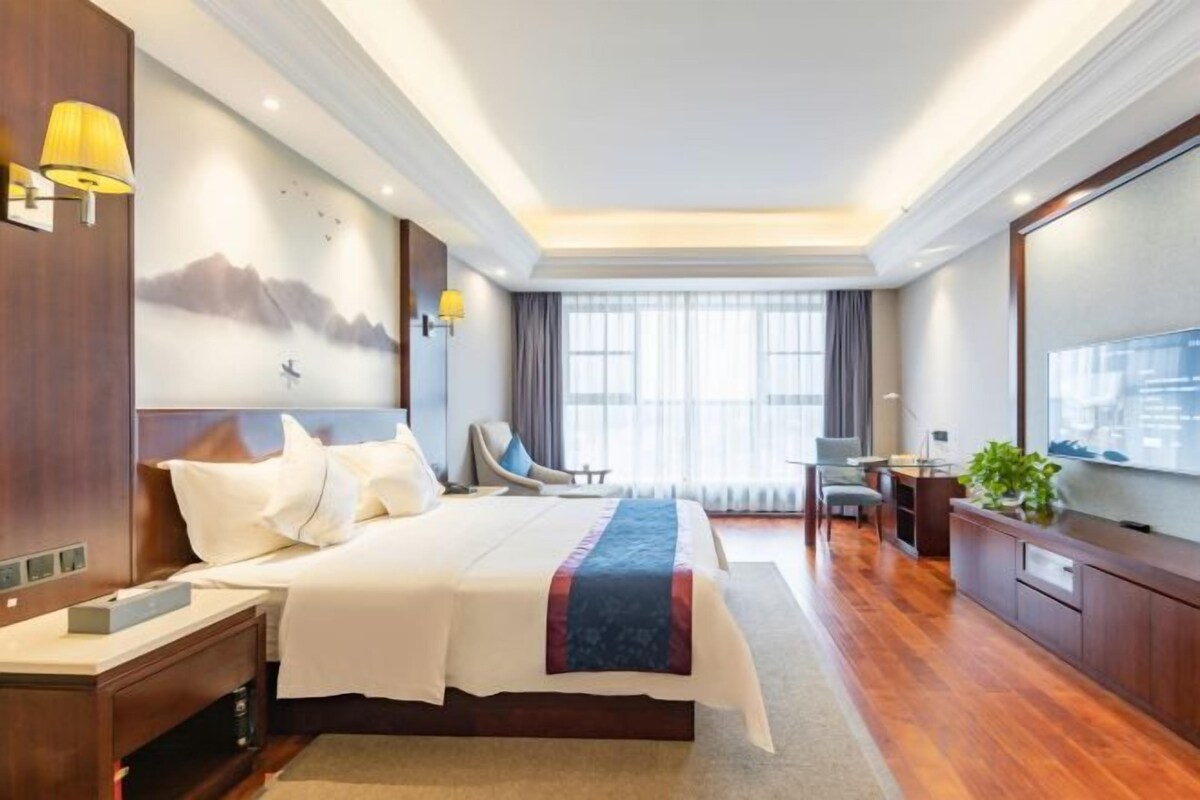 Une chambre d’hôtel avec un grand lit, un bureau, une chaise, une télévision et une fenêtre avec des rideaux.