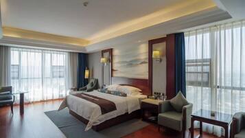 A modern hotel room with a large bed, a desk, a chair, and a large window with curtains.