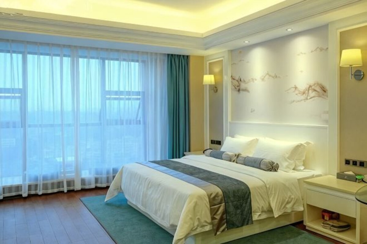 Een hotelkamer met een groot bed, een nachtkastje, gordijnen voor het raam en een muur met een decoratief schilderwerk.