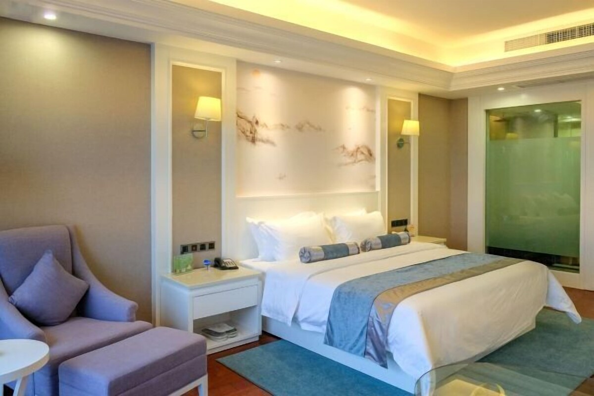 Een hotelkamer met een bed, een stoel, een nachtkastje en een wandlamp.
