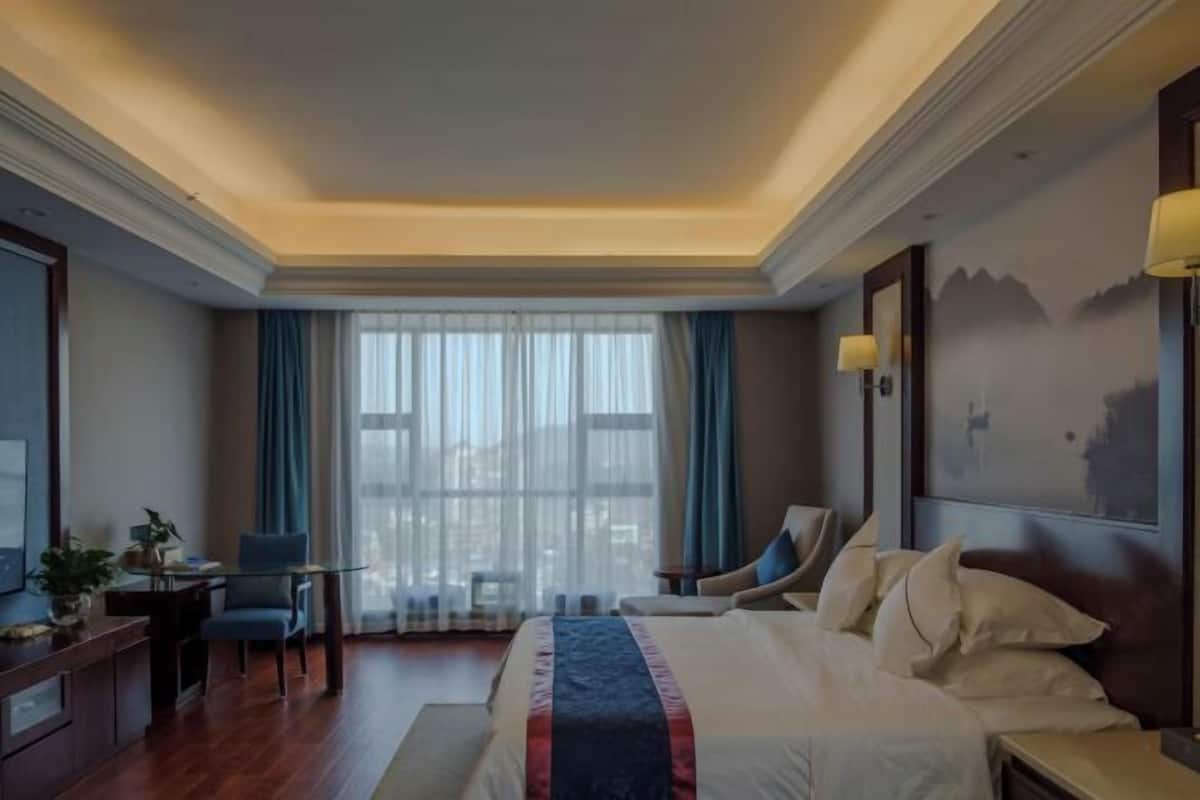 Habitación de hotel con una cama grande, un escritorio con silla, una mesita de noche con lámpara y vistas a la ciudad a través de amplias ventanas.