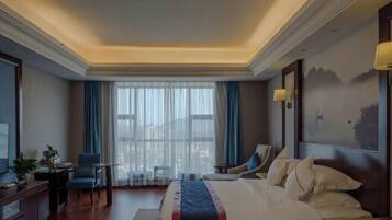 Habitación de hotel con una cama grande, un escritorio con silla, una mesita de noche con lámpara y vistas a la ciudad a través de amplias ventanas.