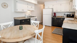A modern kitchen with white cabinets, a gas stove, a refrigerator, and a dining area with a wooden table and white chairs. - Cozy, Roomy Ranch near IU Stadiums (Bloomington)