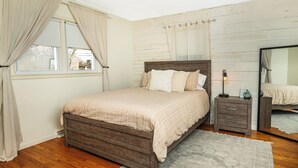 A bedroom with a wooden bed, a nightstand, a bedside lamp, a mirror, and a window with curtains. - Cozy, Roomy Ranch near IU Stadiums (Bloomington)