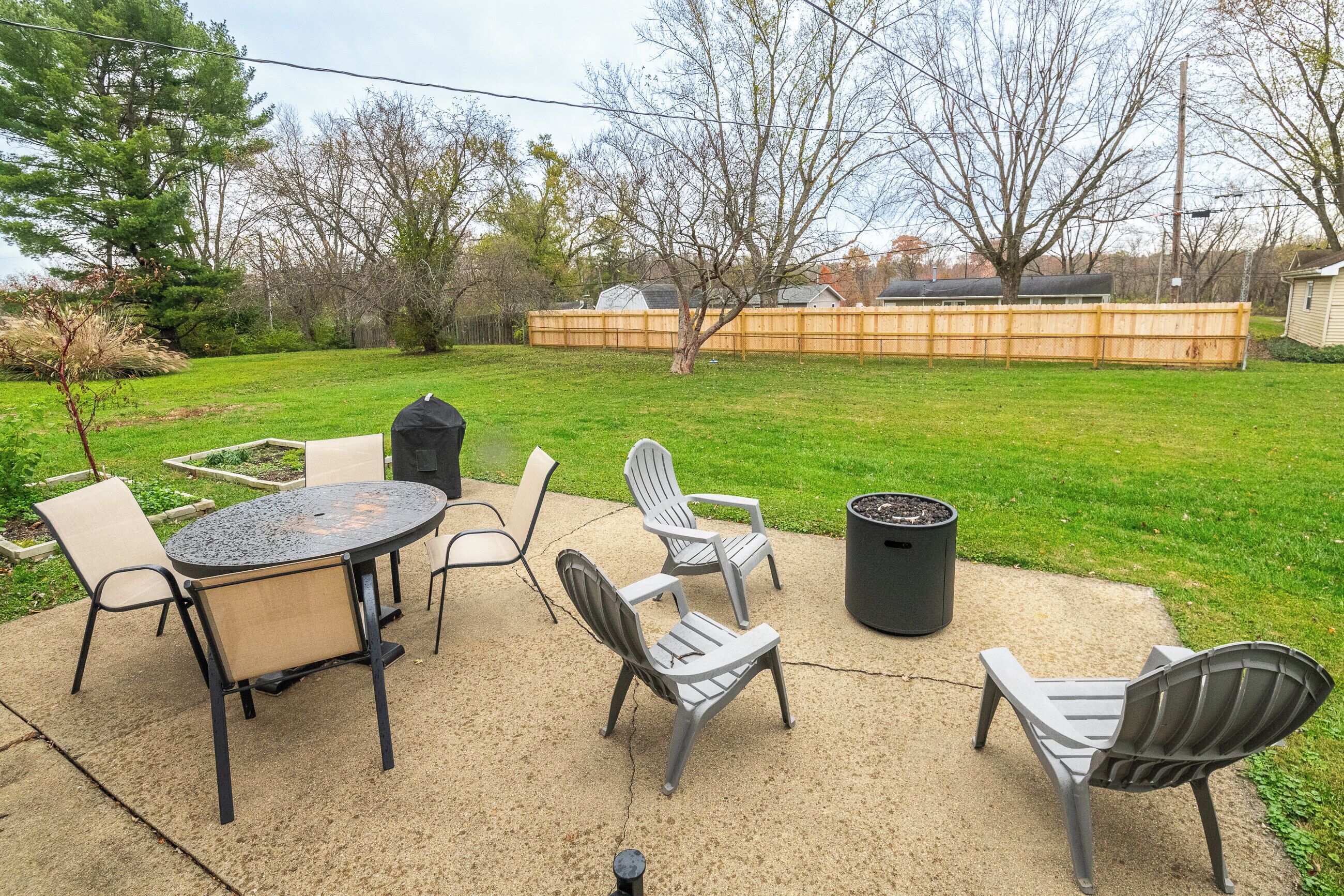 An outdoor patio with a round table and four Adirondack chairs, surrounded by a wooden fence and trees.