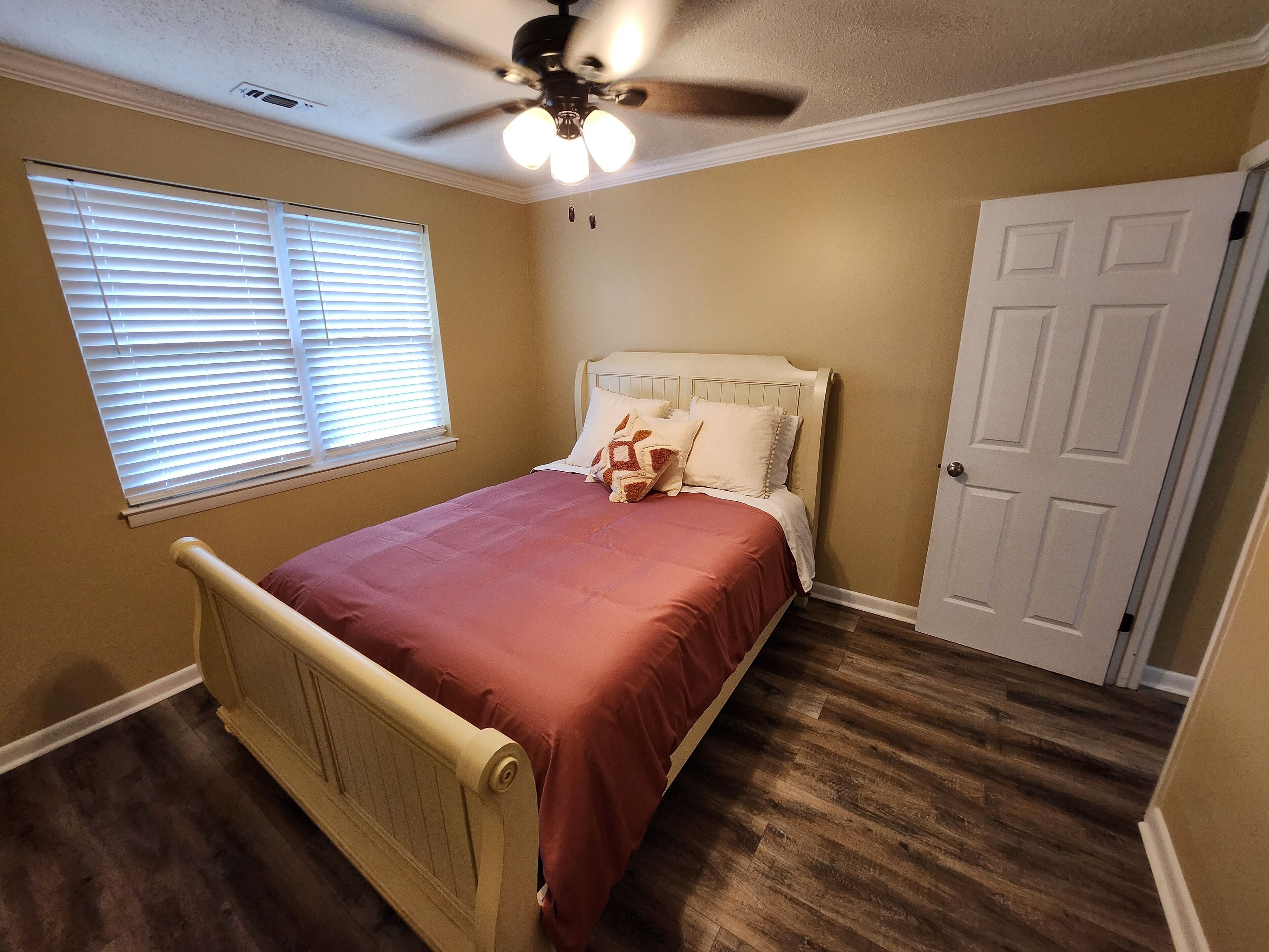 A bedroom with a bed, a ceiling fan, a window with blinds, a door, and wooden flooring.