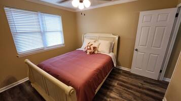 A bedroom with a bed, a ceiling fan, a window with blinds, a door, and wooden flooring.