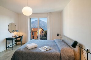 A modern hotel room with a large bed, a desk, a chair, and a view of a mountain and town. - Modern House Apartment 4, Menaggio, Italy (Menaggio)