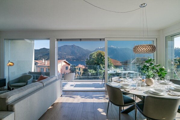 A modern living room with a sofa, a dining area with a table set for four, and a balcony with a mountain view.