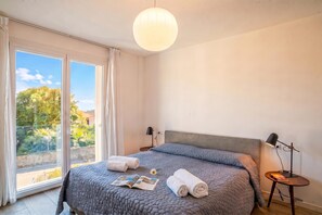 Een slaapkamer met een groot bed, een nachtkastje met een lamp en uitzicht op groen buiten het raam.