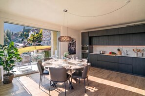 A modern dining area with a large table set for four, a view of a garden through a glass door, and a kitchen with dark cabinetry and a marble countertop.
