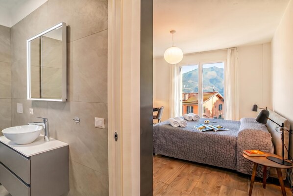 A modern bathroom with a large mirror, a white sink, and a wooden floor. The bedroom features a bed with a patterned bedspread, a bedside table with a lamp, and a view of mountains through the window.