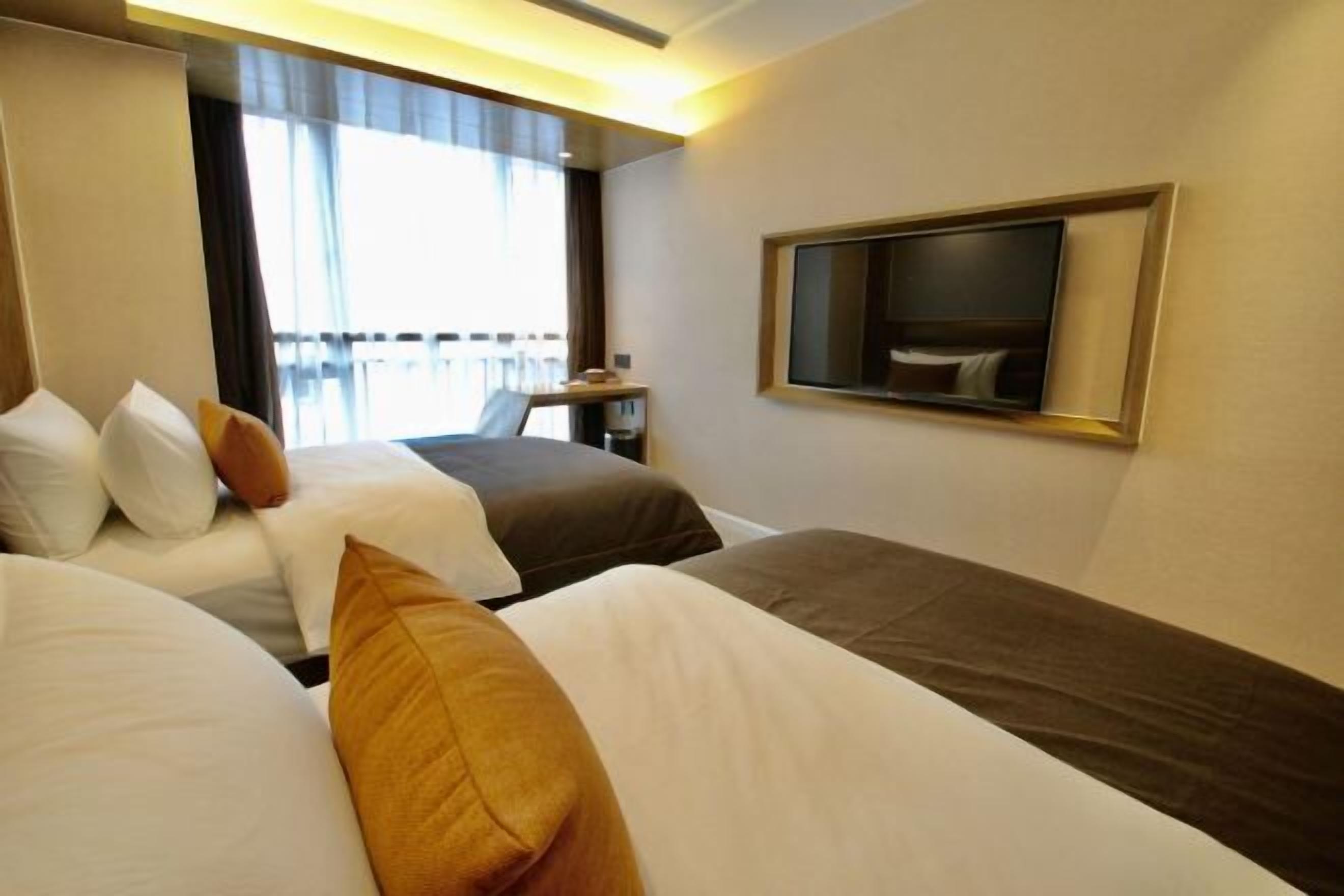 Ein Hotelzimmer mit zwei Betten, einem an der Wand befestigten Fernseher und einem Fenster mit Vorhängen.