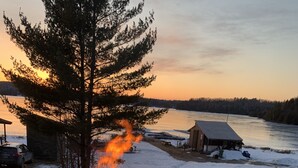 Un feu de camp dans un foyer métallique, avec un paysage enneigé et une rivière en arrière-plan.