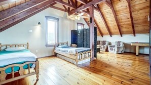 Une chambre spacieuse située dans les combles, avec des poutres apparentes, deux lits, une fenêtre et un ventilateur de plafond.