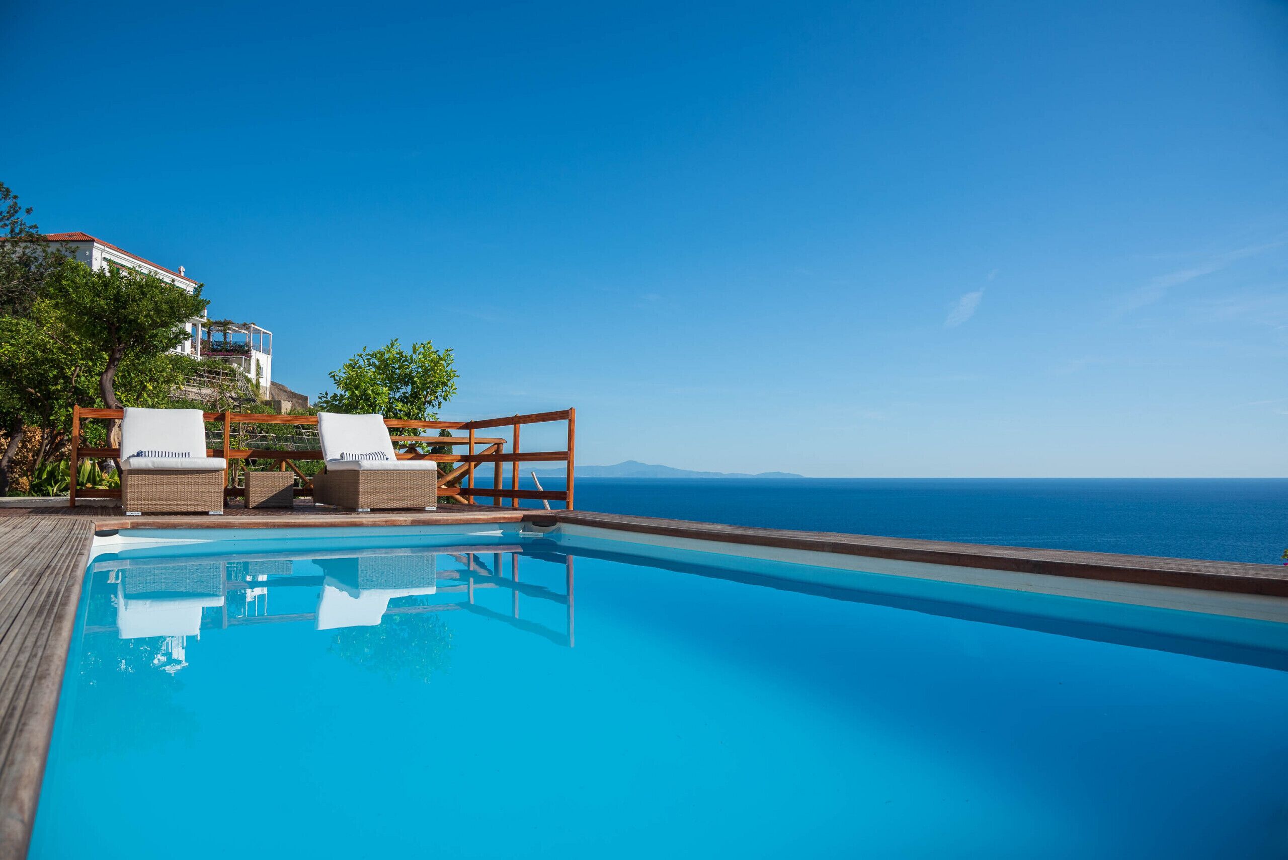 A clear blue pool with lounge chairs and a wooden deck overlooking the sea.