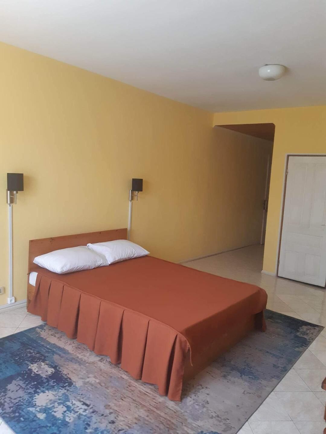 Een slaapkamer met een bed, twee wandlampen, een deur en een kamerkleed met een patroon.