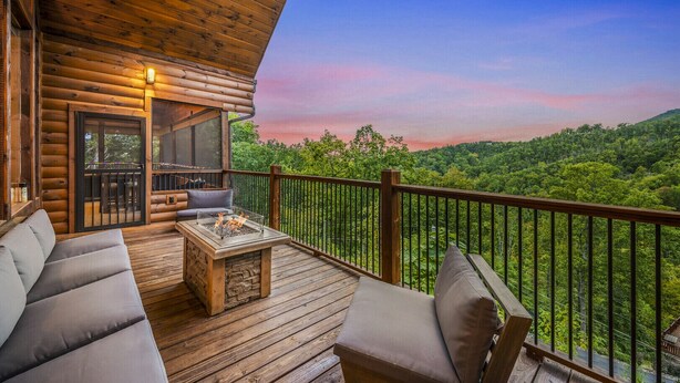 Una terraza de madera con un pozo de fuego, zona de estar y vistas a un paisaje boscoso.