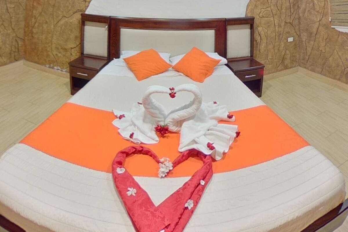 Una habitación de hotel con techo de madera, una cama redonda con ropa de cama naranja, una decoración con forma de corazón y una mesita de noche.