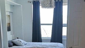 Ein modernes Schlafzimmer mit Bett, kleinem Tisch und Stuhl. Ein Fenster mit Vorhängen ist zu sehen, sowie eine Tür.