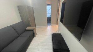 Un salon avec un canapé gris, un pouf noir, une petite table d’appoint en bois et un grand téléviseur à écran plat fixé au mur.