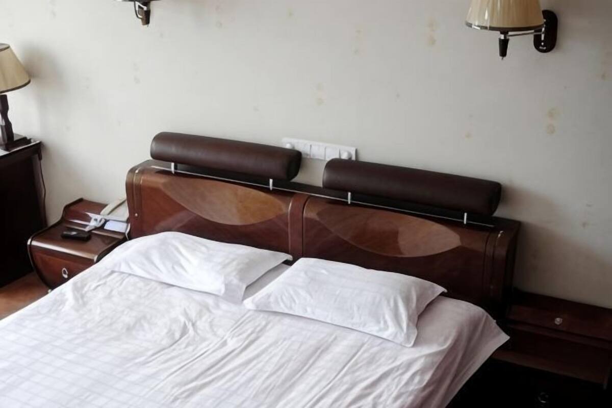 Habitación de hotel con cabecera de madera, cama con sábanas blancas, mesita de noche con lámpara y aplique de pared.