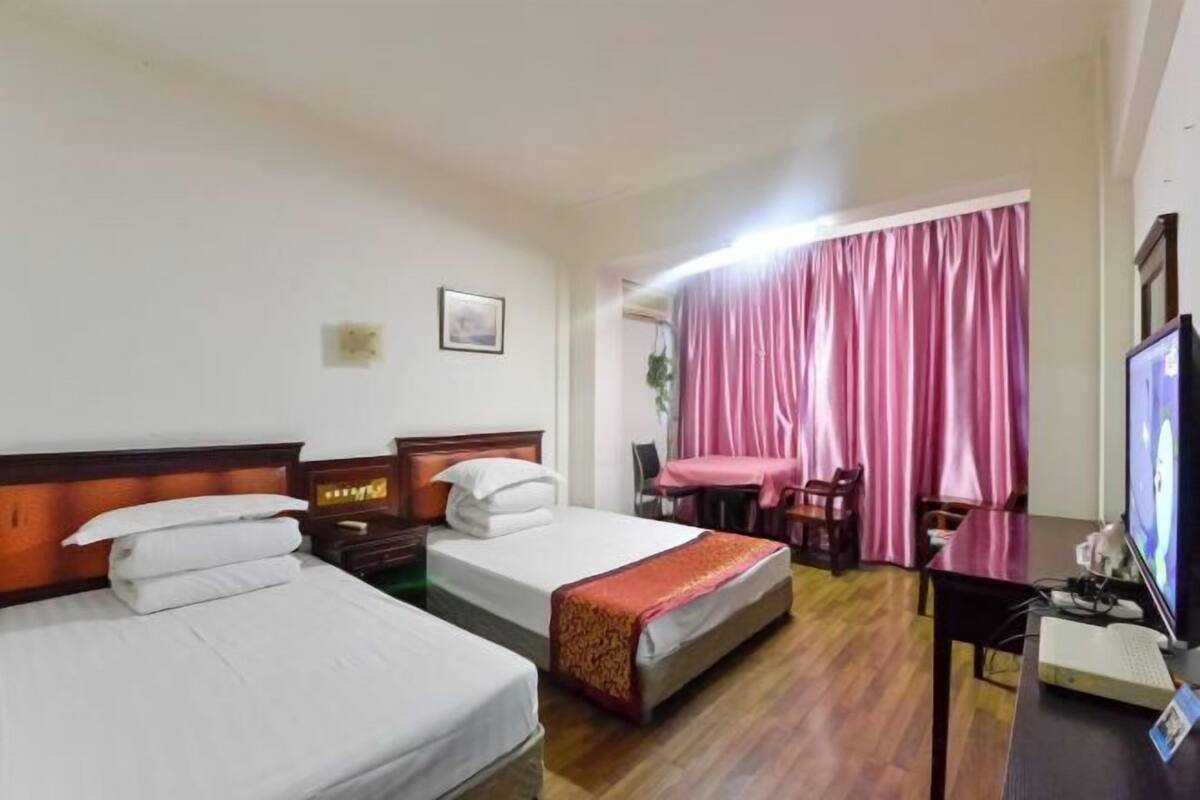 Habitación de hotel con dos camas, un escritorio con computadora, televisión y ventana con cortinas rosas.