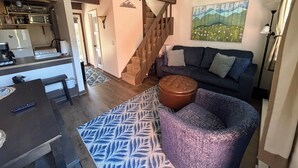 Una sala de estar con un sofá azul oscuro, un tapiz estampado, una escalera de madera y una cocina con microondas y estufa.