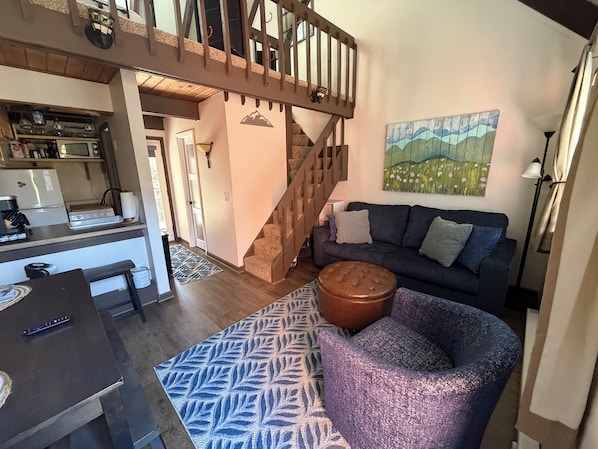 A living room with a dark blue sofa, a patterned rug, a wooden staircase, and a kitchen area with a microwave and stove.